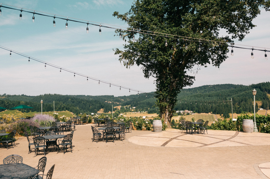 Patio with tables and chairs and an oak tree, overlooking the valley view at Sweet Cheeks Winery.
