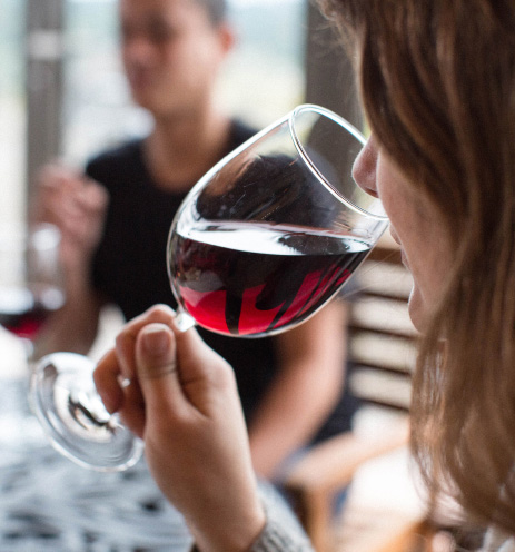 Woman smelling a glass of red wine, man in the background with a glass of red wine.