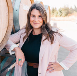 Photo of General Manager, Jessica Thomas, leaning arm on barrels of wine.
