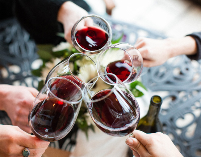 Five hands cheering glasses of red wine together over a floral arrangement.
