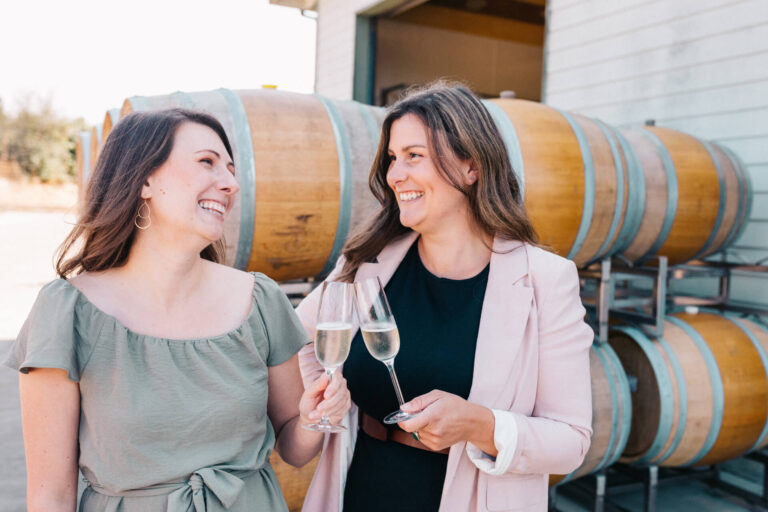 Two women cheering flutes of sparkling wine with barrels of wine in the background.